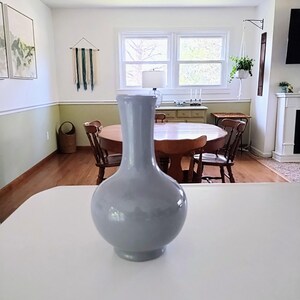 May include: A light gray ceramic vase with a rounded body and a long, slender neck. The vase is placed on a white countertop, with a dining room and window in the background. The vase is a decorative item.