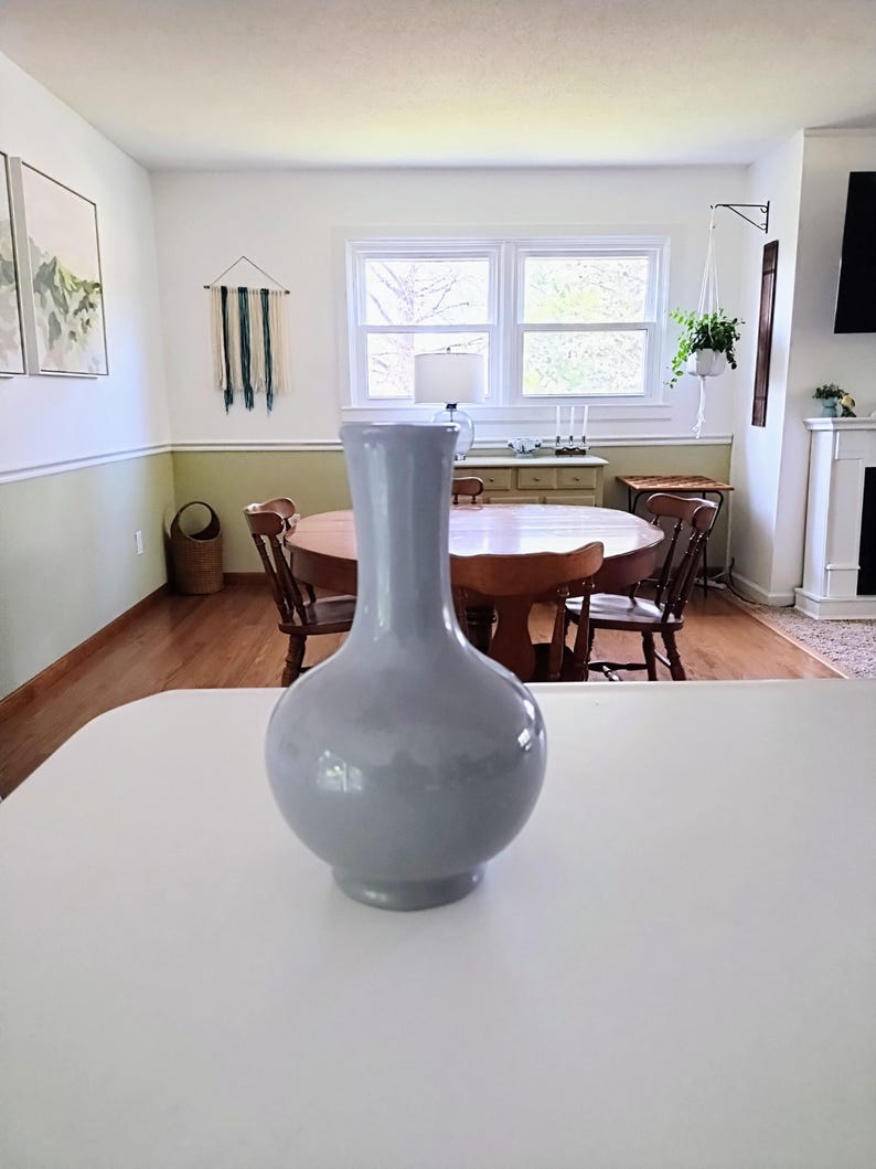 May include: A light gray ceramic vase with a rounded body and a long, slender neck. The vase is placed on a white countertop, with a dining room and window in the background. The vase is a decorative item.
