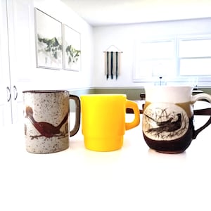 May include: Three ceramic mugs on a white surface. The mugs are different colors and designs. One is yellow, one is speckled with a bird design, and the other is brown and white with a bird and landscape design.