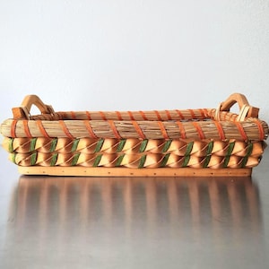 May include: A rectangular woven basket featuring a light wood base and two integrated handles. The sides are decorated with a braided pattern of tan and green strips, topped with a rim wrapped in natural fibers and a bright orange cord.