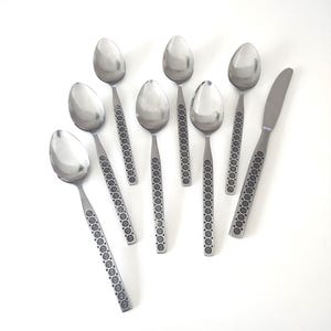 May include: A set of stainless steel flatware, including seven spoons and one knife. The handles feature a repeating floral pattern. The spoons have oval bowls, and the knife has a straight blade.