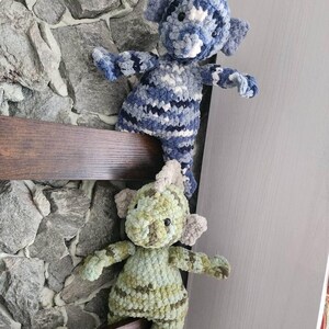 May include: Two handmade crochet stuffed animals in the shape of a dragon or dinosaur. One is blue and white, the other is green and brown. They are positioned on a wooden ledge against a stone wall.