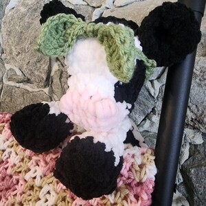 May include: A handmade crochet cow plushie with black and white markings, a pink nose, and green bow-shaped ears. The cow is resting on a pink, white, and tan crochet blanket. The plushie is soft and cuddly.