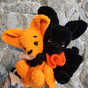 May include: Two plush bunny toys. One is orange with black accents and the other is black with an orange bow tie. The toys have large ears and are made of a soft, textured material. The background is a gray stone wall.