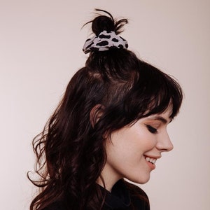May include: A woman with long, dark brown hair styled in a messy bun with a black and white leopard print scrunchie. She is wearing a black turtleneck sweater and is smiling.