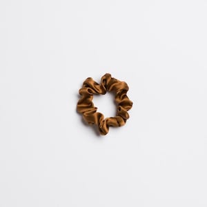 May include: A single, scrunchie in a rich, warm brown color. The hair accessory is made of a silky material, with a gathered, ruffled design. The scrunchie is circular and sits against a white background.