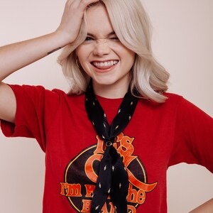 以下が含まれることがあります： 「I'm with the band」と書かれたグラフィックの赤いTシャツと、稲妻模様の黒いスカーフ。スカーフは円形のリングで固定されています。その人はTシャツとスカーフを着ています。