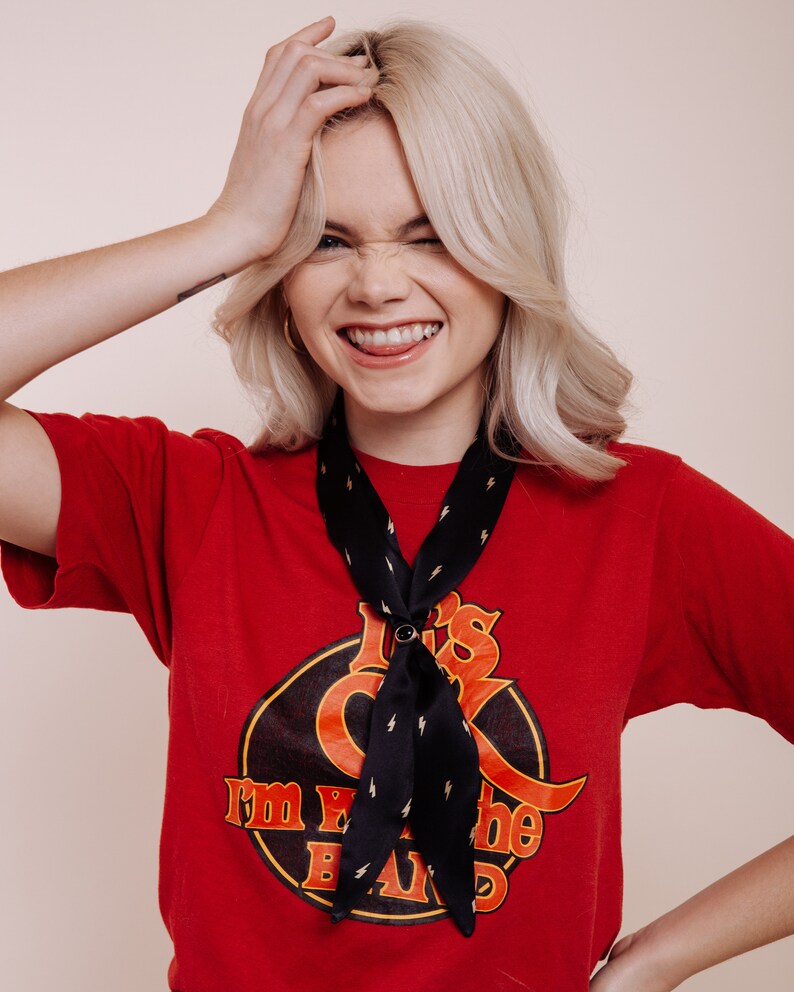以下が含まれることがあります： 「I'm with the band」と書かれたグラフィックの赤いTシャツと、稲妻模様の黒いスカーフ。スカーフは円形のリングで固定されています。その人はTシャツとスカーフを着ています。