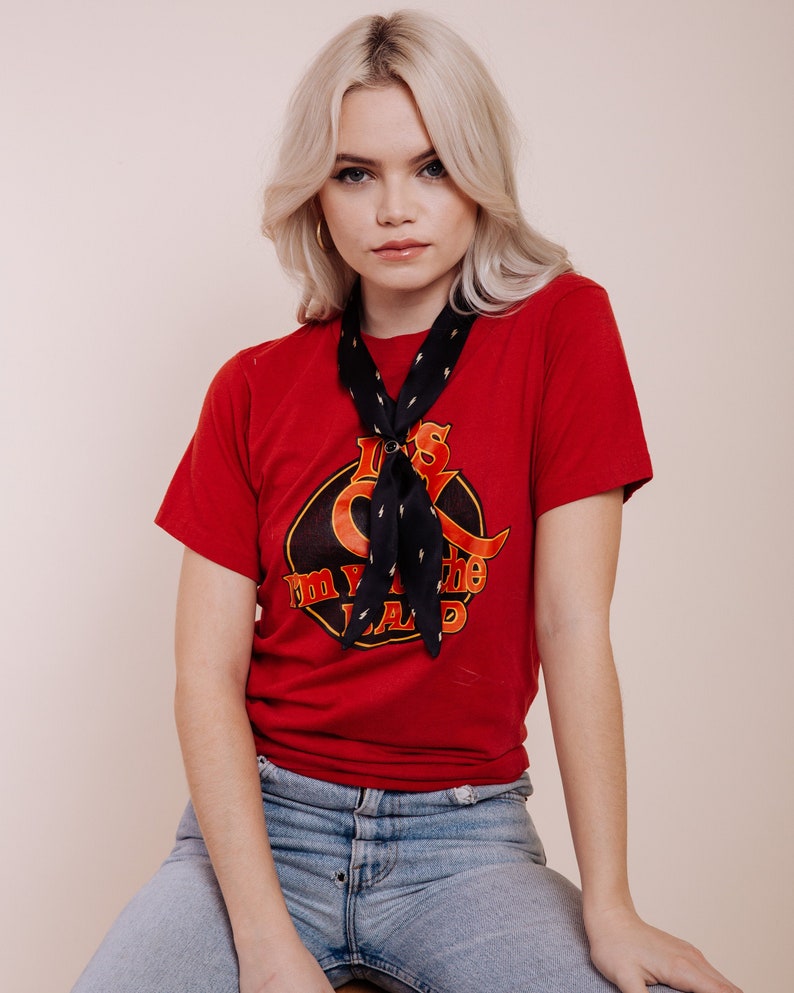 以下が含まれることがあります： 「I'm in the band」の文字とグラフィックデザインが施された赤いTシャツ。白い稲妻模様の黒いスカーフが首に巻かれています。青いジーンズを履いています。