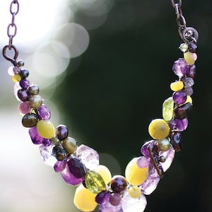 May include: A handcrafted necklace featuring a variety of gemstones and pearls. The necklace showcases a mix of purple amethyst, yellow beads, green stones, and dark pearls, strung on a silver chain. The design is organic and eye-catching.
