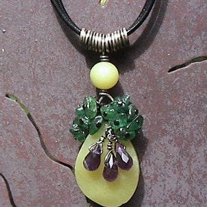May include: A necklace with a pear-shaped pendant made of yellow stone and green and purple gemstones. The pendant is surrounded by green gemstones.