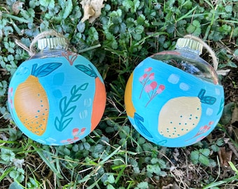 May include: Two hand-painted glass ornaments with a blue background and a lemon design. One ornament features a yellow lemon with a green leaf and a pink flower. The other ornament features a yellow lemon with a green leaf and a pink flower.