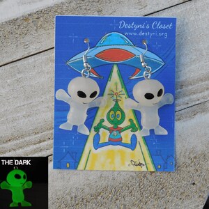 May include: Alien-themed earrings featuring two small, translucent alien figures with black eyes and outstretched arms. The earrings are displayed on a blue card with an illustration of a UFO and other aliens. The card reads "Destyni's Closet".