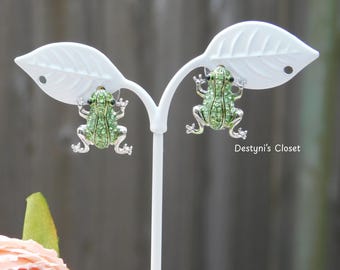 May include: A pair of frog-shaped stud earrings. The earrings are green with silver-colored accents and small clear crystals. The frog's body is covered in small green crystals. The earrings are displayed on a white leaf-shaped earring holder.