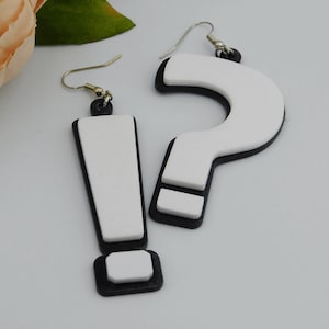 May include: A pair of novelty earrings, one shaped like an exclamation point and the other a question mark. The earrings are white with black borders and silver-colored hooks. The earrings are a fun, playful accessory.