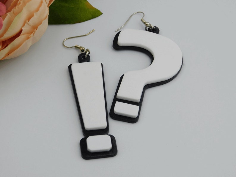 May include: A pair of novelty earrings, one shaped like an exclamation point and the other a question mark. The earrings are white with black borders and silver-colored hooks. The earrings are a fun, playful accessory.