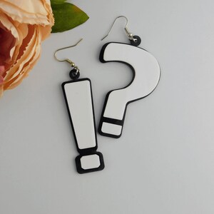 May include: A pair of novelty earrings featuring a white exclamation point and a white question mark, both outlined in black. The earrings hang from silver-colored hooks and black circular accents. The background is a light gray.