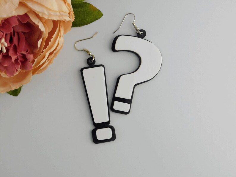 May include: A pair of novelty earrings featuring a white exclamation point and a white question mark, both outlined in black. The earrings hang from silver-colored hooks and black circular accents. The background is a light gray.