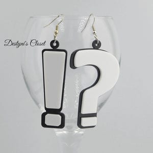 May include: A pair of novelty earrings featuring an exclamation point and a question mark. The earrings are white with black outlines and black tops. The earrings are hanging from silver-colored hooks. The text "Destyni's Closet" is visible in the background.