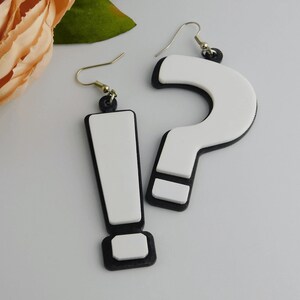 May include: A pair of novelty earrings featuring a white exclamation point and a white question mark, both outlined in black. The earrings have gold-colored hooks and black accents at the top. The background includes a soft pink flower and green leaves.