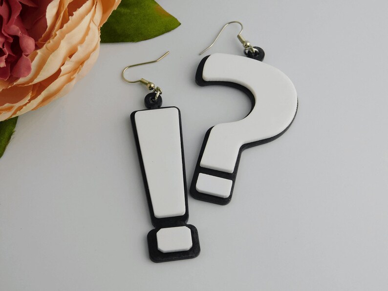 May include: A pair of novelty earrings featuring a white exclamation point and a white question mark, both outlined in black. The earrings have gold-colored hooks and black accents at the top. The background includes a soft pink flower and green leaves.