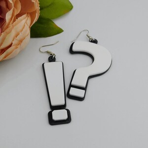 May include: A pair of quirky earrings featuring an exclamation point and a question mark. The earrings have a white center with a black border and gold-colored hooks. The exclamation point earring is rectangular, and the question mark earring is curved.