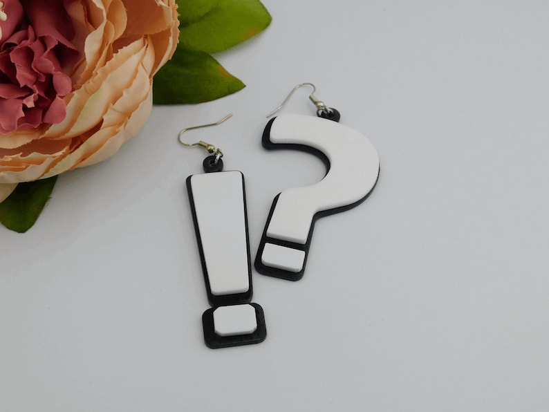 May include: A pair of quirky earrings featuring an exclamation point and a question mark. The earrings have a white center with a black border and gold-colored hooks. The exclamation point earring is rectangular, and the question mark earring is curved.