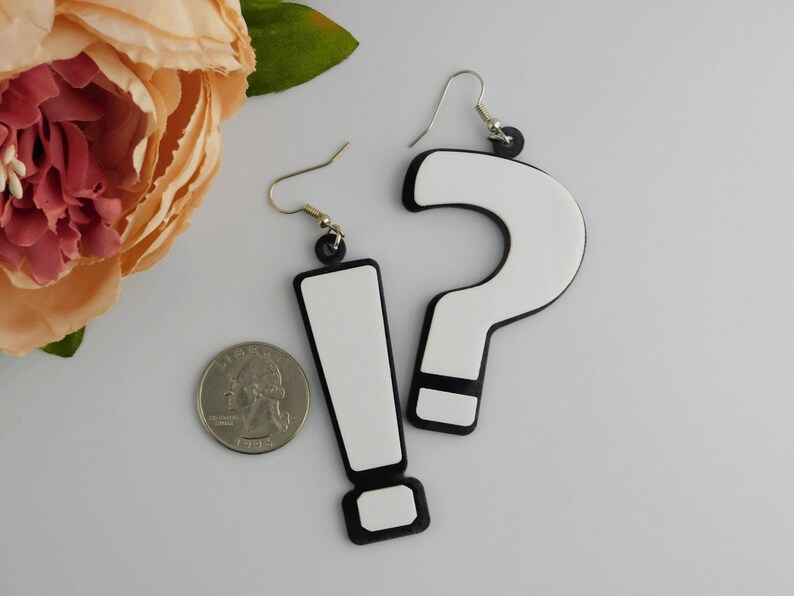 May include: A pair of novelty earrings featuring a white exclamation point and a white question mark, both outlined in black. The earrings have silver-colored hooks and are displayed on a light gray surface.