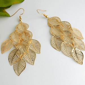 May include: A pair of gold-colored leaf earrings. Each earring features multiple layered, openwork leaves suspended from a hook. The leaves have detailed veining, and the earrings are displayed on a white surface.
