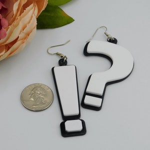 May include: A pair of novelty earrings featuring an exclamation point and a question mark. The earrings are white with black trim and silver-colored hooks. A U.S. quarter is next to the earrings for size comparison.