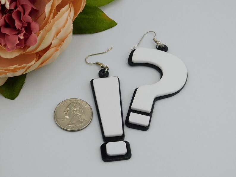 May include: A pair of novelty earrings featuring an exclamation point and a question mark. The earrings are white with black trim and silver-colored hooks. A U.S. quarter is next to the earrings for size comparison.