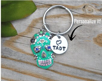 May include: A teal sugar skull keychain with silver accents and a small, round tag. The tag has a heart and the initials "TADT". The keychain is on a silver ring. The text "Personalize it!" is in the background.