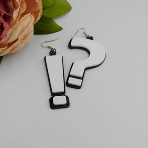 May include: A pair of novelty earrings, one shaped like an exclamation point and the other like a question mark. The earrings are white with black outlines and accents, and have silver-colored ear wires. The earrings are likely made of acrylic or a similar material.