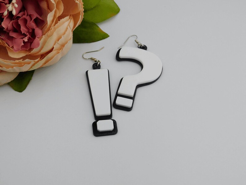 May include: A pair of novelty earrings, one shaped like an exclamation point and the other like a question mark. The earrings are white with black outlines and accents, and have silver-colored ear wires. The earrings are likely made of acrylic or a similar material.