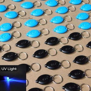 May include: An assortment of blue and black UV LED keychain lights with silver key rings. The lights are oval-shaped with a small button. The image includes a smaller inset showing the UV light in action.