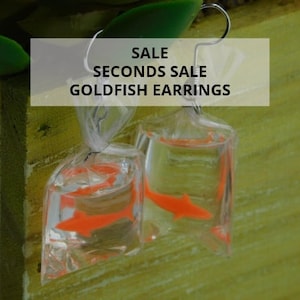 May include: Goldfish earrings in clear plastic bags with orange fish inside. The earrings have silver hooks. The text on the image says "SALE SECONDS SALE GOLDFISH EARRINGS."