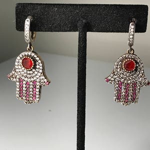 Puede incluir: Un par de pendientes de plata con un diseño de mano de hamsa. Los pendientes están adornados con piedras de circonita cúbica transparentes y rojas.