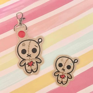 May include: Tan voodoo doll keychain and patch set. The keychain has a silver clasp and a red accent. The voodoo dolls have black eyes, stitched details, and a red heart. The background is a colorful striped pattern.