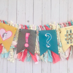 以下が含まれることがあります： ユニコーン、感嘆符、疑問符、ハッシュタグが描かれたカラフルなバナー。バナーは布の切れ端で作られており、素朴で手作り感のある外観です。