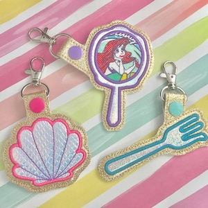 Puede incluir: Tres llaveros de color dorado con diferentes diseños. El primer llavero presenta una concha con escamas iridiscentes. El segundo llavero presenta un tenedor dorado con un mango turquesa. El tercer llavero presenta un espejo con un mango morado y una imagen de Ariel de La Sirenita.
