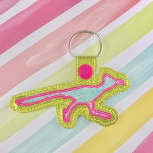 May include: A green glitter keychain with a pink outline of a bird with a floral pattern inside. The keychain has a silver ring and a pink circle on the top.