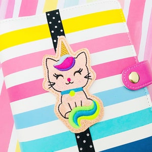 May include: A whimsical cat-unicorn applique in pastel colors, with a rainbow mane and tail, and a golden horn, is attached to a striped planner. The planner has a pink snap closure and a black ribbon with white polka dots.