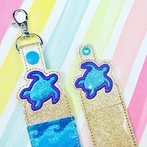 May include: Two gold glitter keychains with a blue and purple sea turtle design. The keychains are on a striped background.