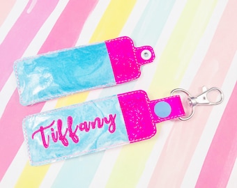 Puede incluir: Un llavero de tela rosa y azul con el nombre "Tiffany" bordado en rosa. El llavero tiene un anillo de metal plateado y un círculo azul.
