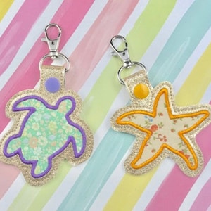 May include: Two keychains with a floral fabric design. One keychain features a purple outlined sea turtle and the other features a yellow outlined starfish.