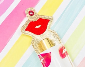 Puede incluir: Un porta-labios de tela blanca con ribete de purpurina dorada y estampado de labios rojos. El porta-labios tiene un diseño de labios de purpurina dorada en la parte superior con un agujero para un llavero.