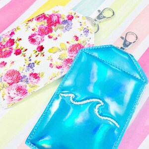 May include: Two keychains with different designs. One keychain is white with a floral pattern and the other is a bright blue iridescent keychain with a white wave design.