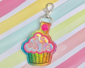 Può includere: Un portachiavi a forma di cupcake arcobaleno iridescente con un bottone rosa e una chiusura in metallo argentato.