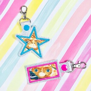 May include: Two keychains featuring a character from the movie E.T. The Extra-Terrestrial. One keychain is a blue star with a silver glitter background. The other keychain is a silver rectangle with a pink glitter background. Both keychains have a silver metal ring.