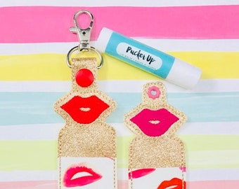 Puede incluir: Dos porta-bálsamos labiales blancos con un acabado de purpurina dorada y un diseño de labios rojos. Los porta-bálsamos tienen un fondo blanco con labios rojos impresos en ellos. Los porta-bálsamos están unidos a un llavero plateado. También se incluye un tubo de bálsamo labial con el texto "Pucker Up".